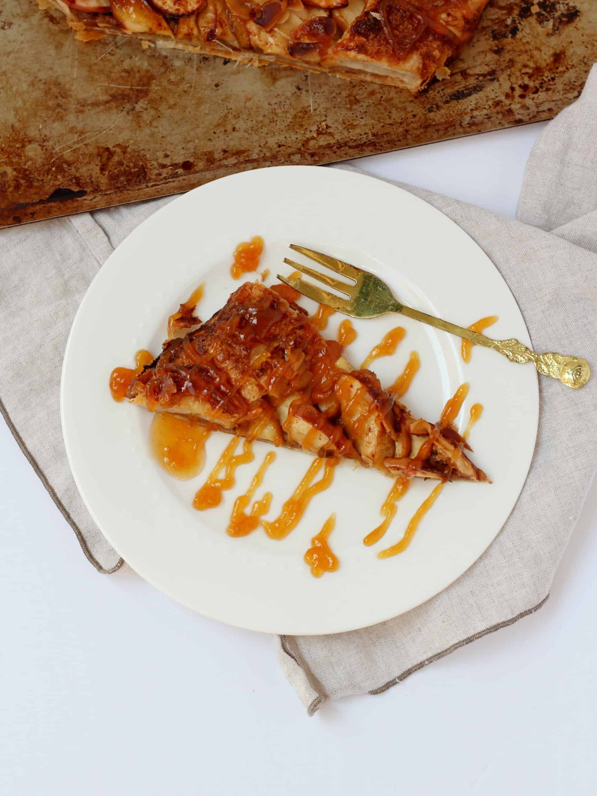 close up of apple galette slice drizzled with caramel sauce on white plate with golden fork next to it
