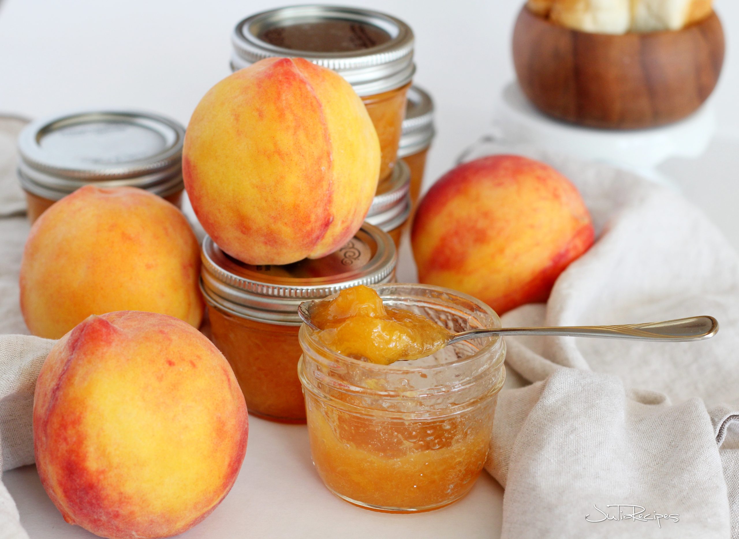 table filled with jars of peach jam, on top of some jars are fresh large peaches, one jar is open and peach jam is spooned out
