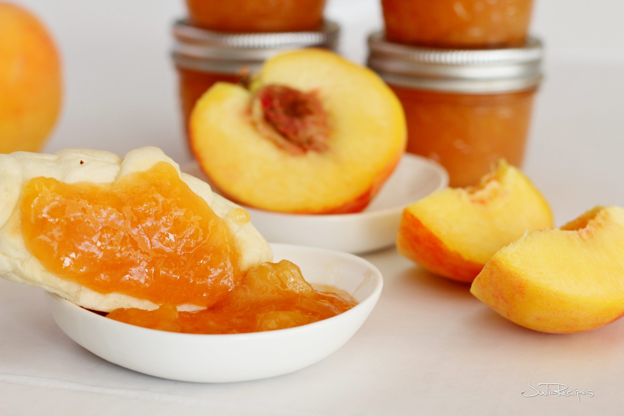 small amount of peach jam in little shallow bowl, right to the left is laying mini croissant spread with the peach jam, along are few cut fresh peaches and peach jam jars 