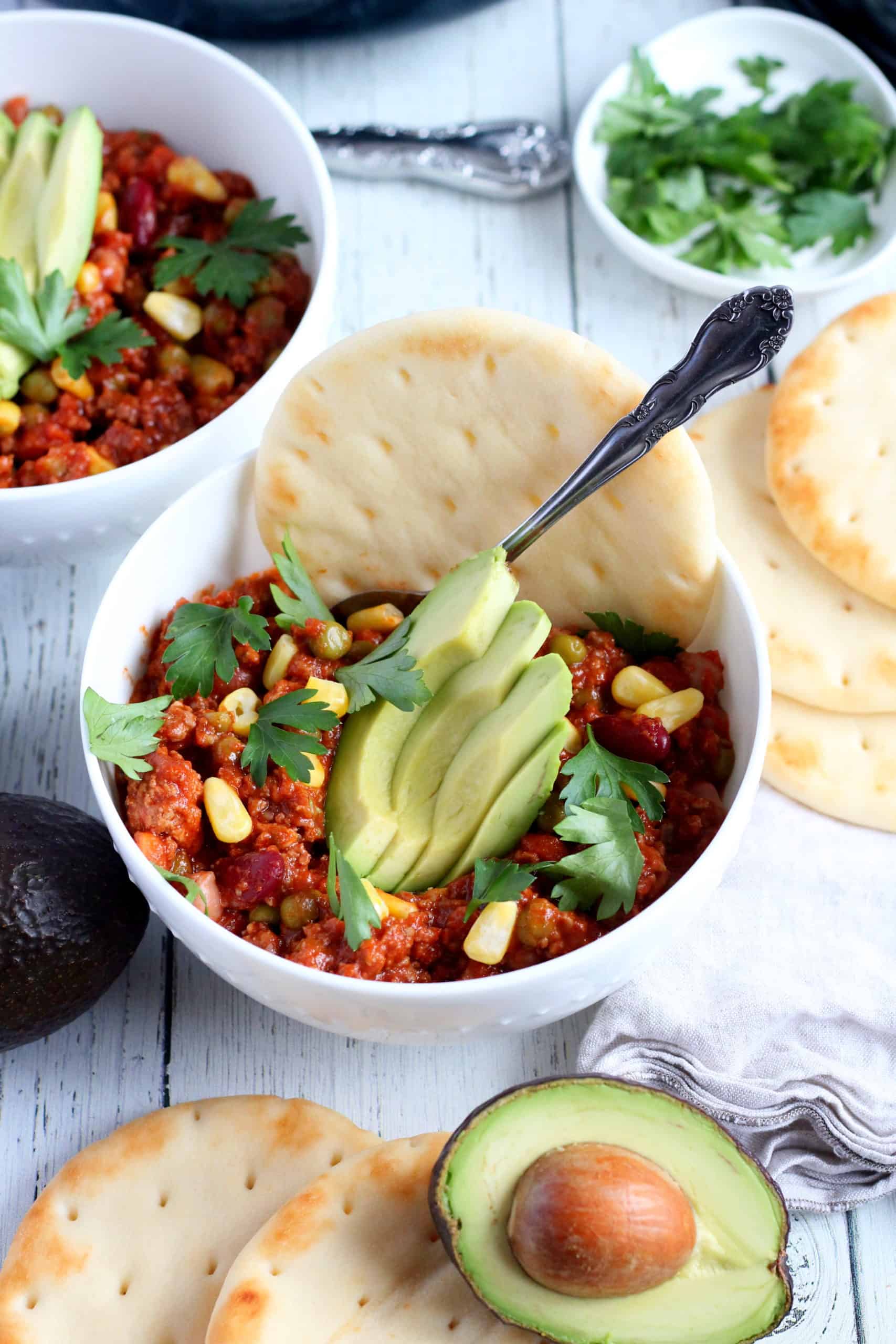 minced meat chili con carne in white bowl topped with avocado and flat bread