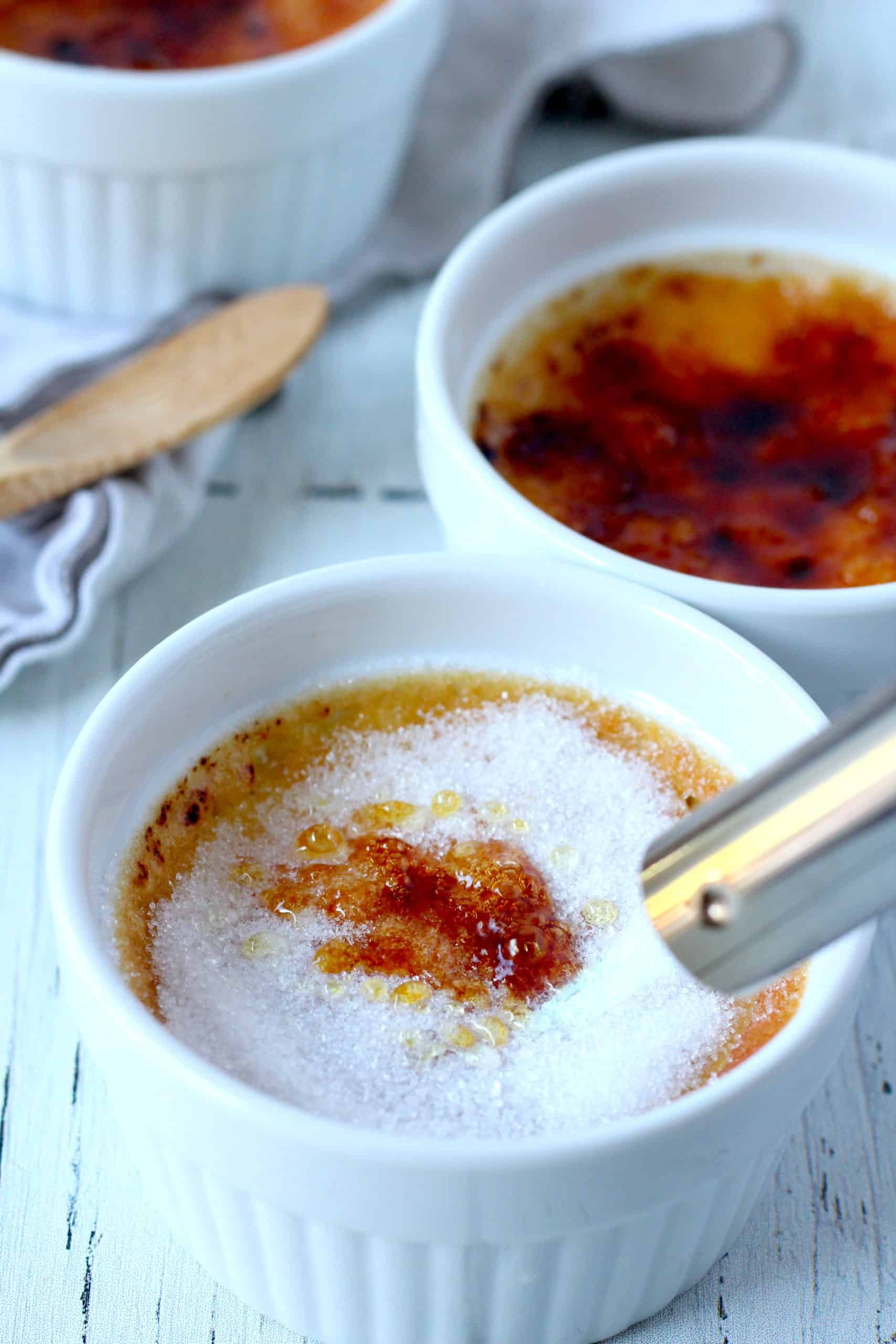 creamy creme brule in white ramekin with sugar being melted by torch another ramekin with creme brulee in background