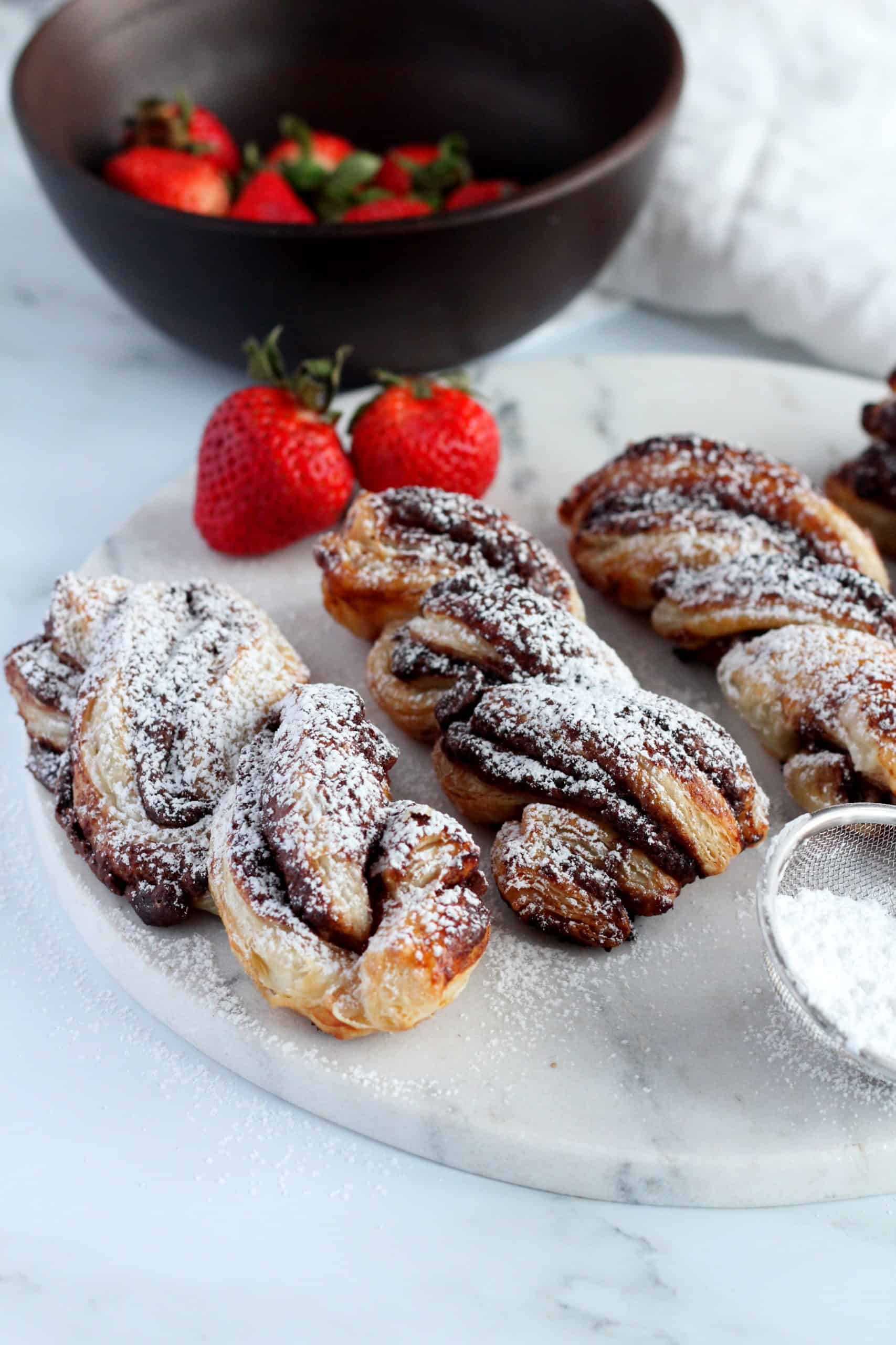Nutella filled puff pastry twists on marble plate dusted with powdered sugar. In the background are strawberries in wooden bowl
