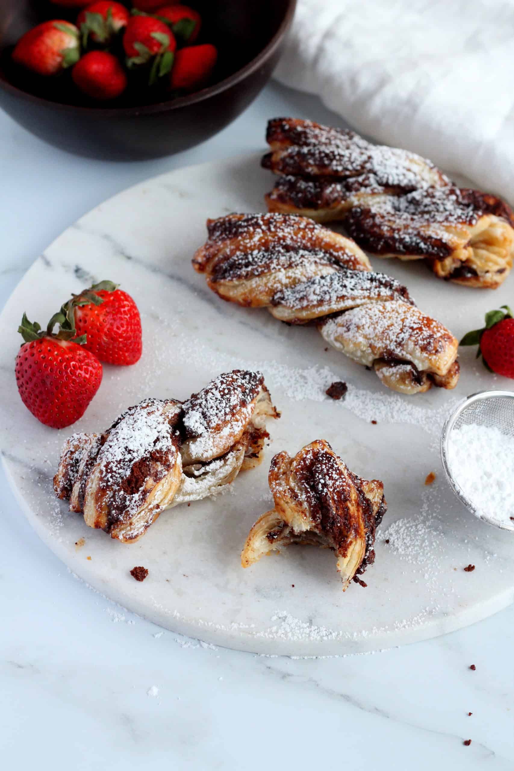 Nutella puff pastry twists on a marble plate with strawberries in the background and powdered sugar wand.