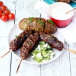 bbq'd kebabs placed on white plate accompanied with cucumber salad and rosted potato in the background glasses with wine cherry tomato vine and tzaziki sauce