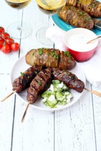 bbq'd kebabs placed on white plate accompanied with cucumber salad and rosted potato in the background glasses with wine cherry tomato vine and tzaziki sauce
