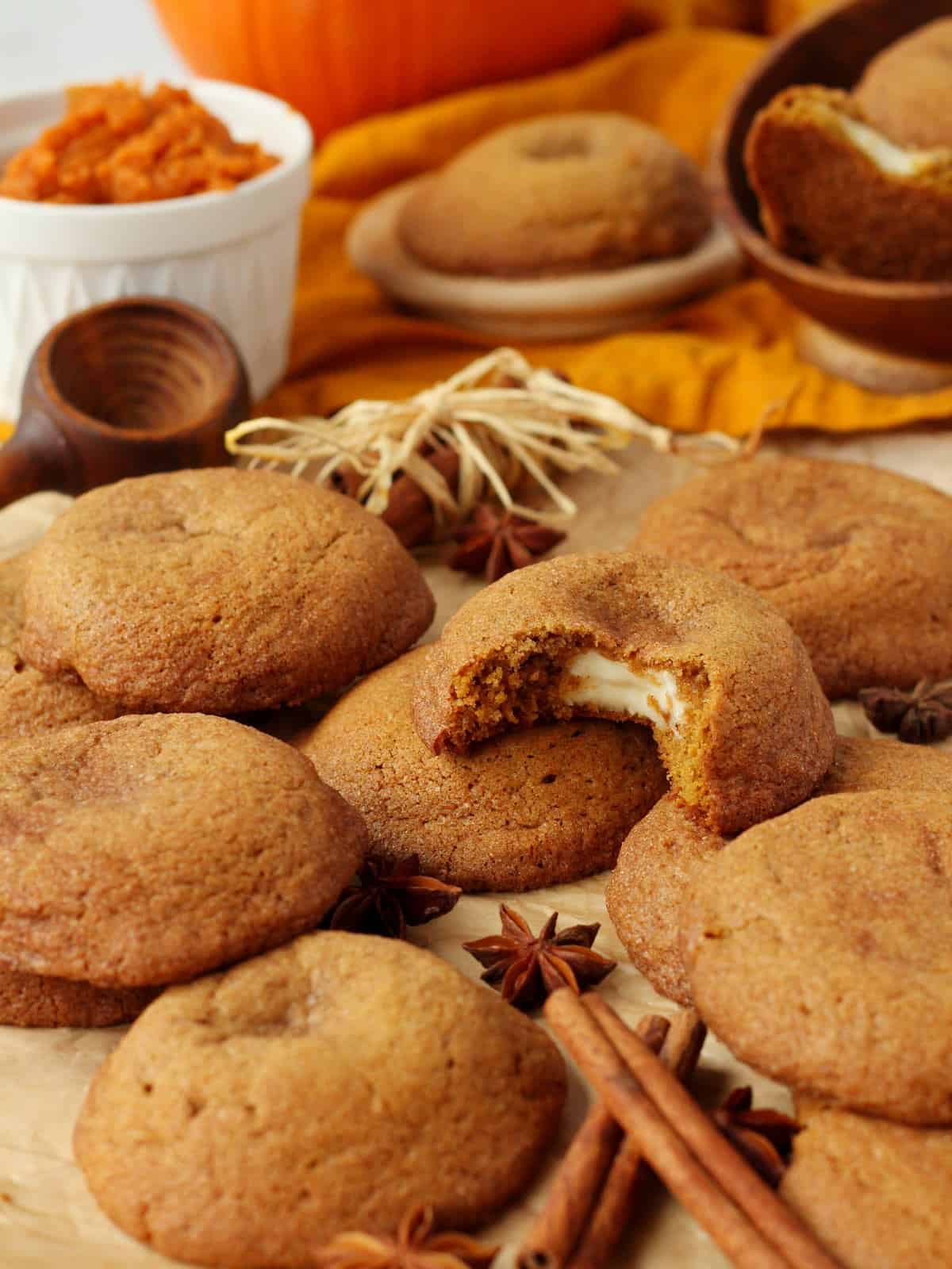 Pumpkin cheesecake cookies scattered on brown parchment paper with pumpkin pure & cinnamon sticks in background