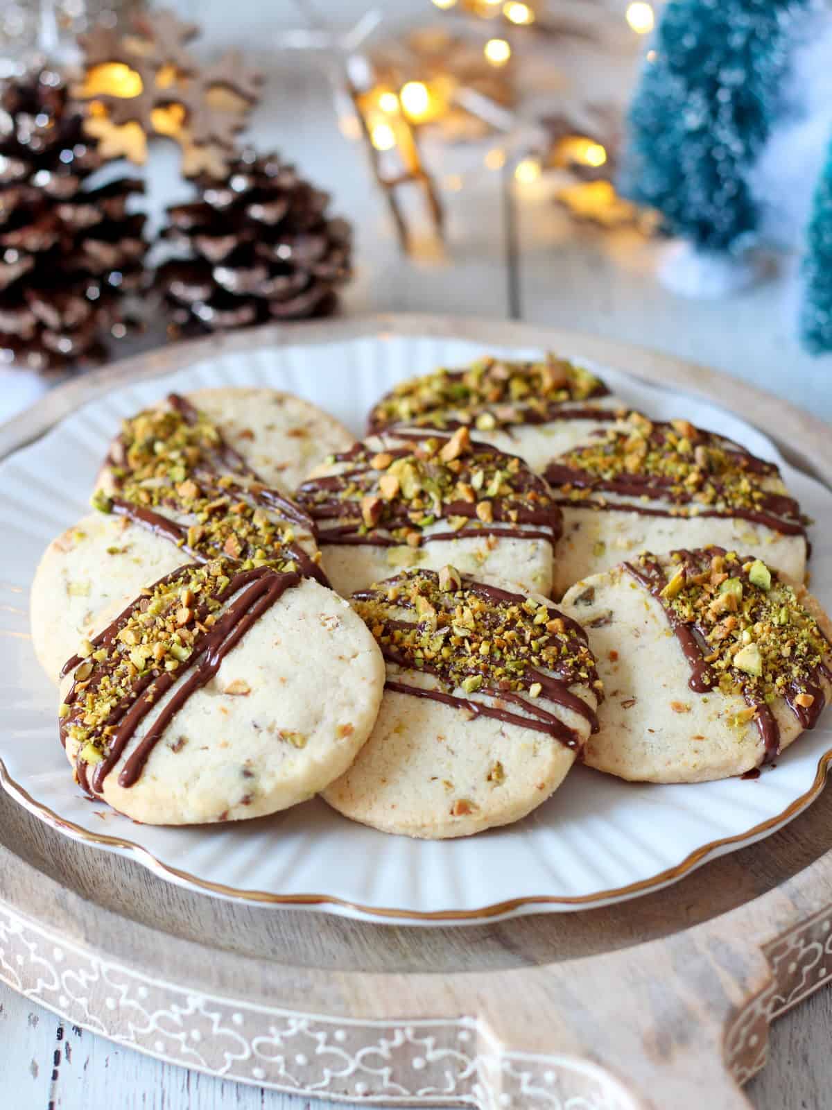 pistachio shortbread cookies drizzled with milk chocolate and placed on white plate with Christmas decoration in background.