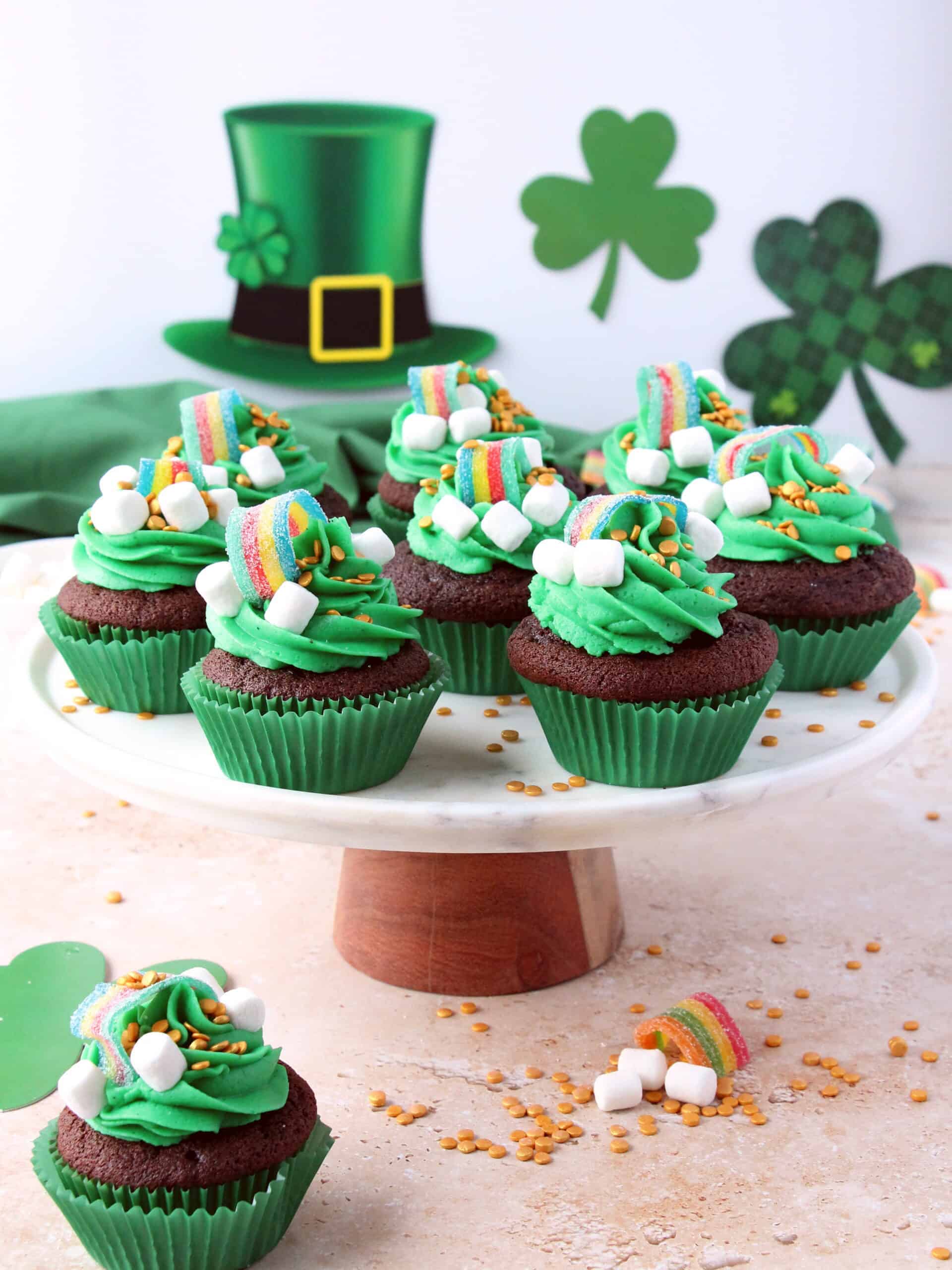st patrick's day cupcakes with green buttercream and raninbow candies on marble cake stand, green decoration in background