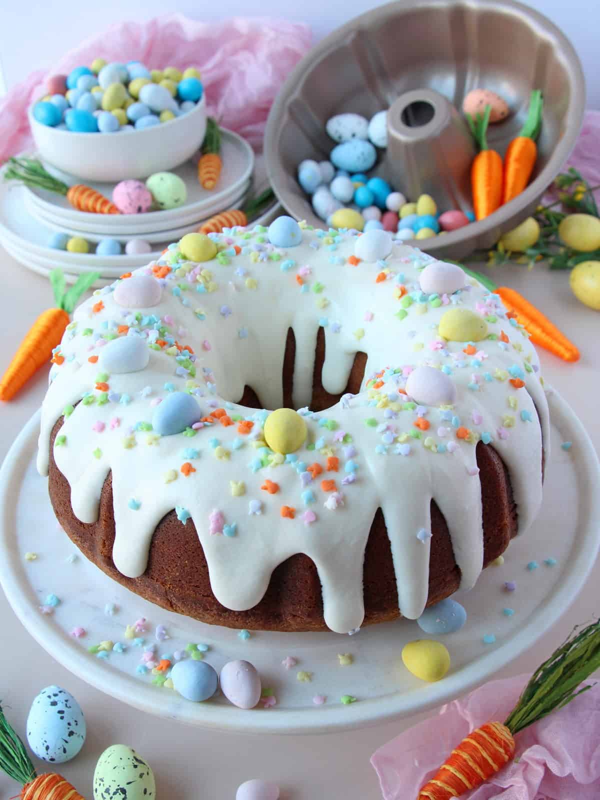 Easter bundt cake glazed with cream cheese and topped with chocolate hershey eggies. Behind plates filled with eggies and colourful easter decoration