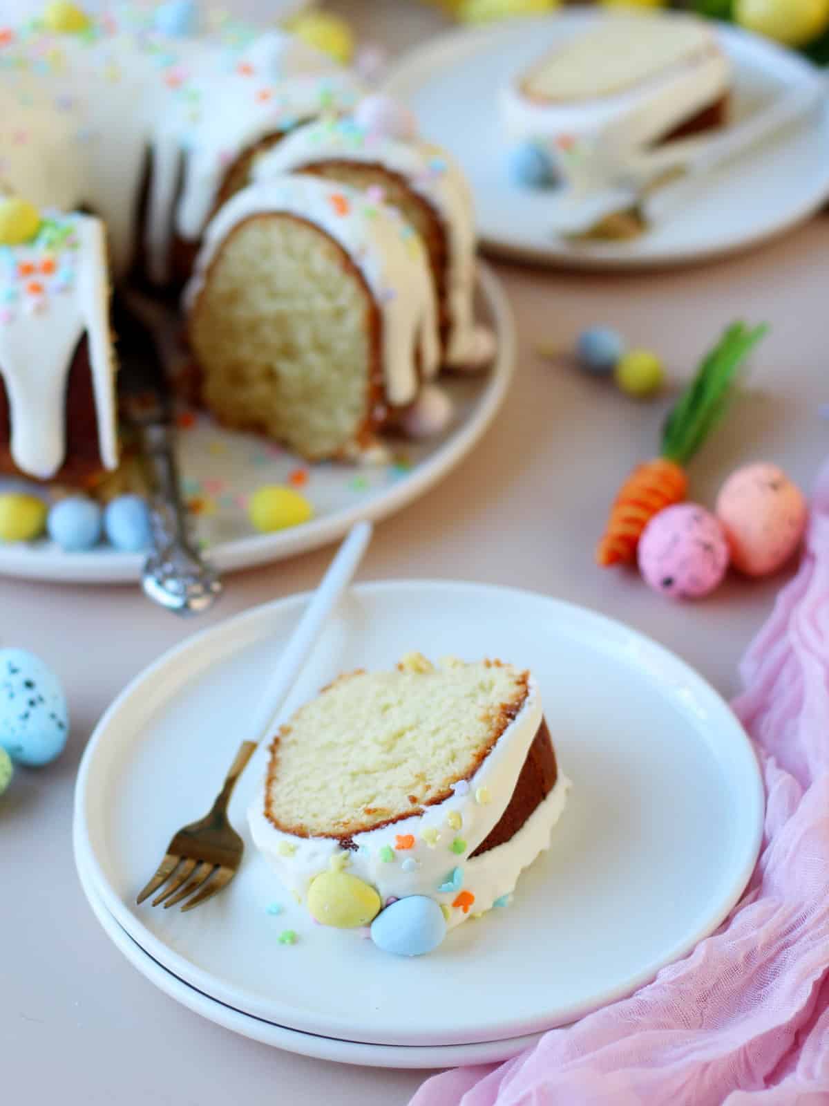 slice of vanilla bundt cake topped with cream cheese and colourful spring sprinkles, the slice of cake is on white plate, behind is the bundt cake with few slices cut on a plate.